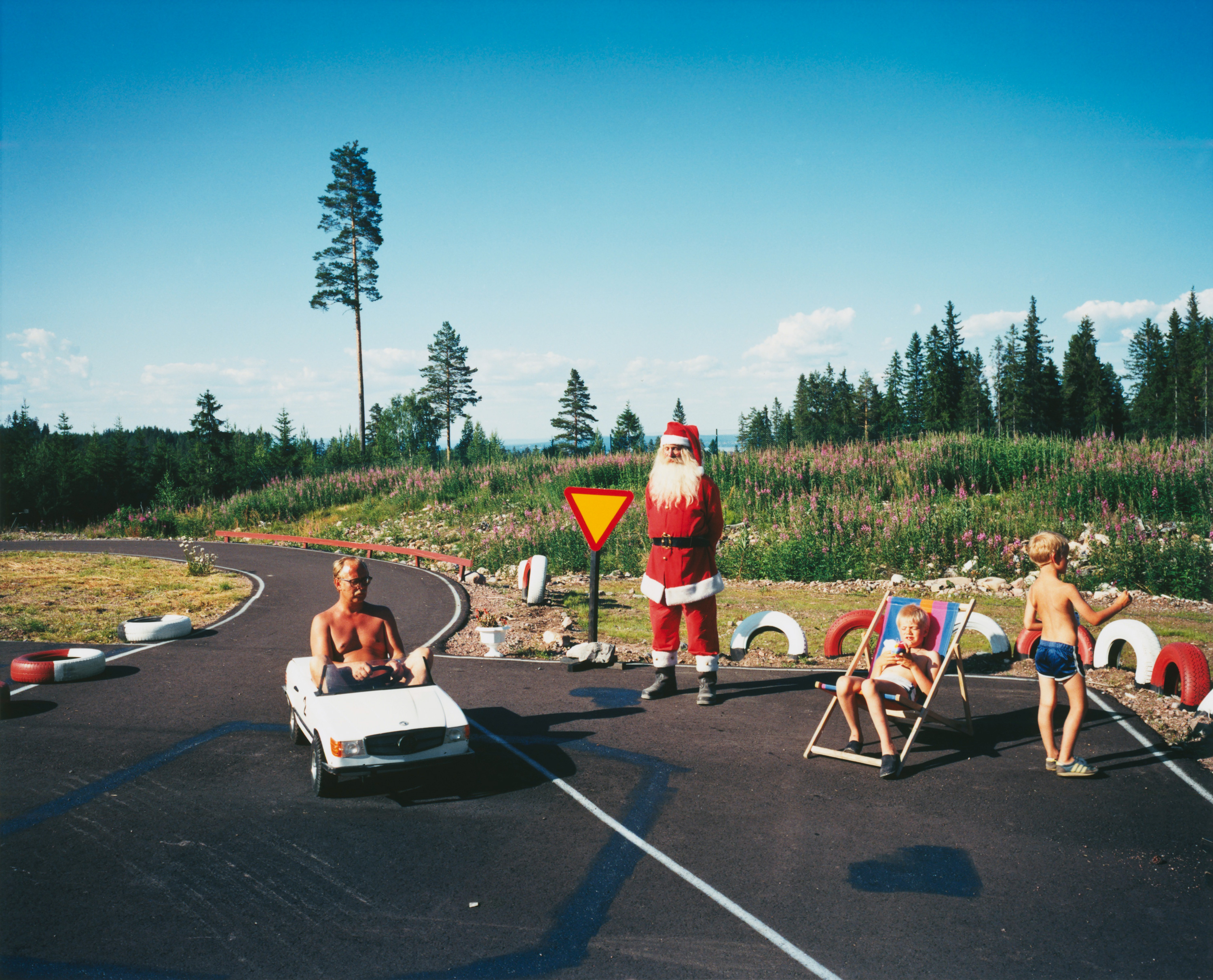 foto av en sommardag vid ett kalhygge och en gokartbana, en man utklädd till tomte står bredvid några uttråkade barn och en man sitter i en leksaksbil och kör runt på banan.