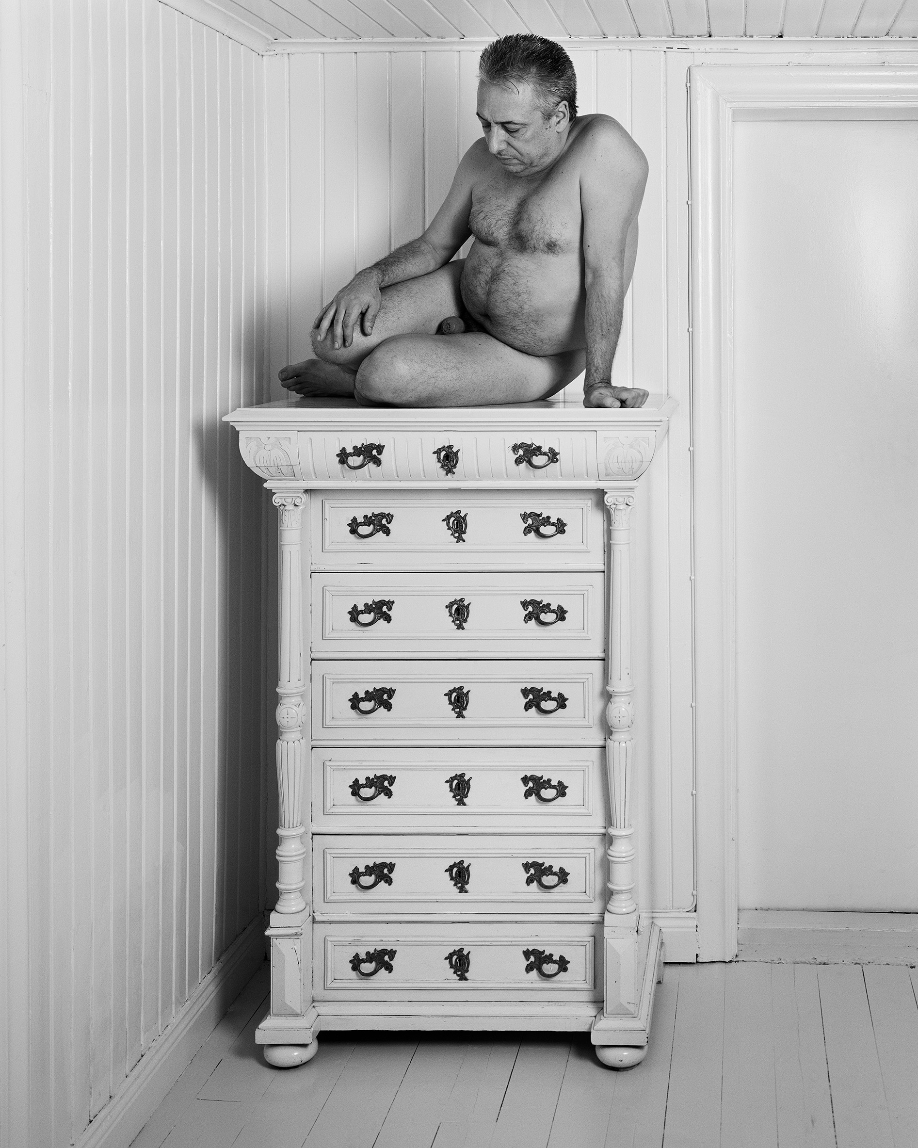 foto i svart vit föreställande en man som sitter naken på en vitmålad byrå