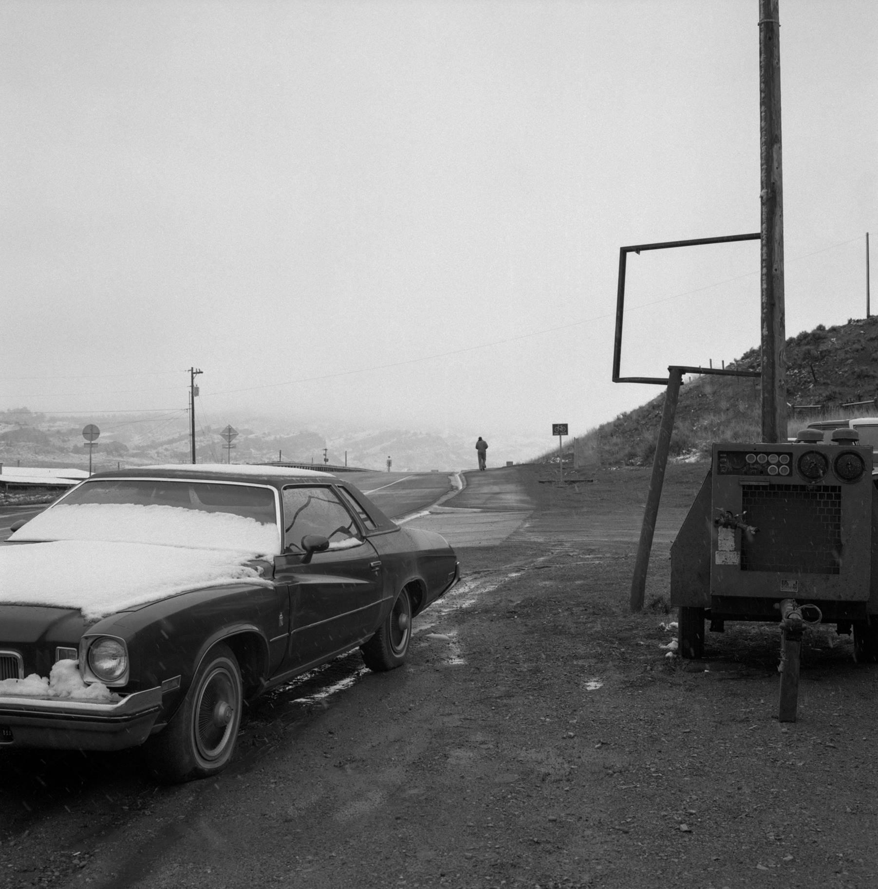 svart vit fotografi med en snötäckt bil i förgrunden och en människogestalt i bakgrunden i slutet av en landsväg