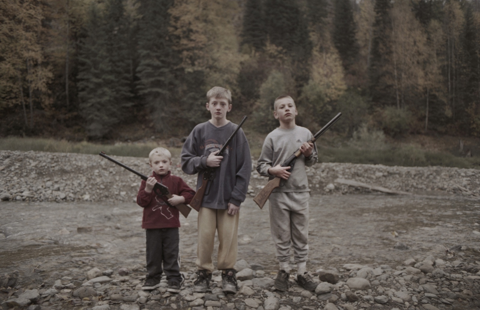 Tre pojkar ca 7-11 år står framför ett vattendrag med gevär i händerna