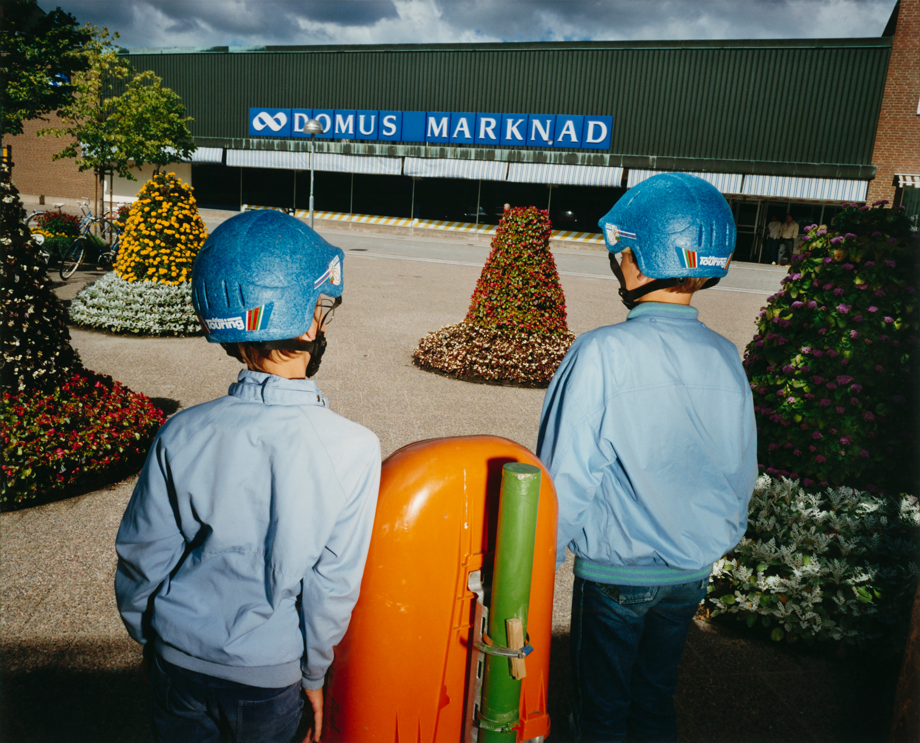 fotografi med två barn i blå cykelhjälmar i förgrunden som blickar ner mot en Domus-butik. Foto av Lars Tunbjörk
