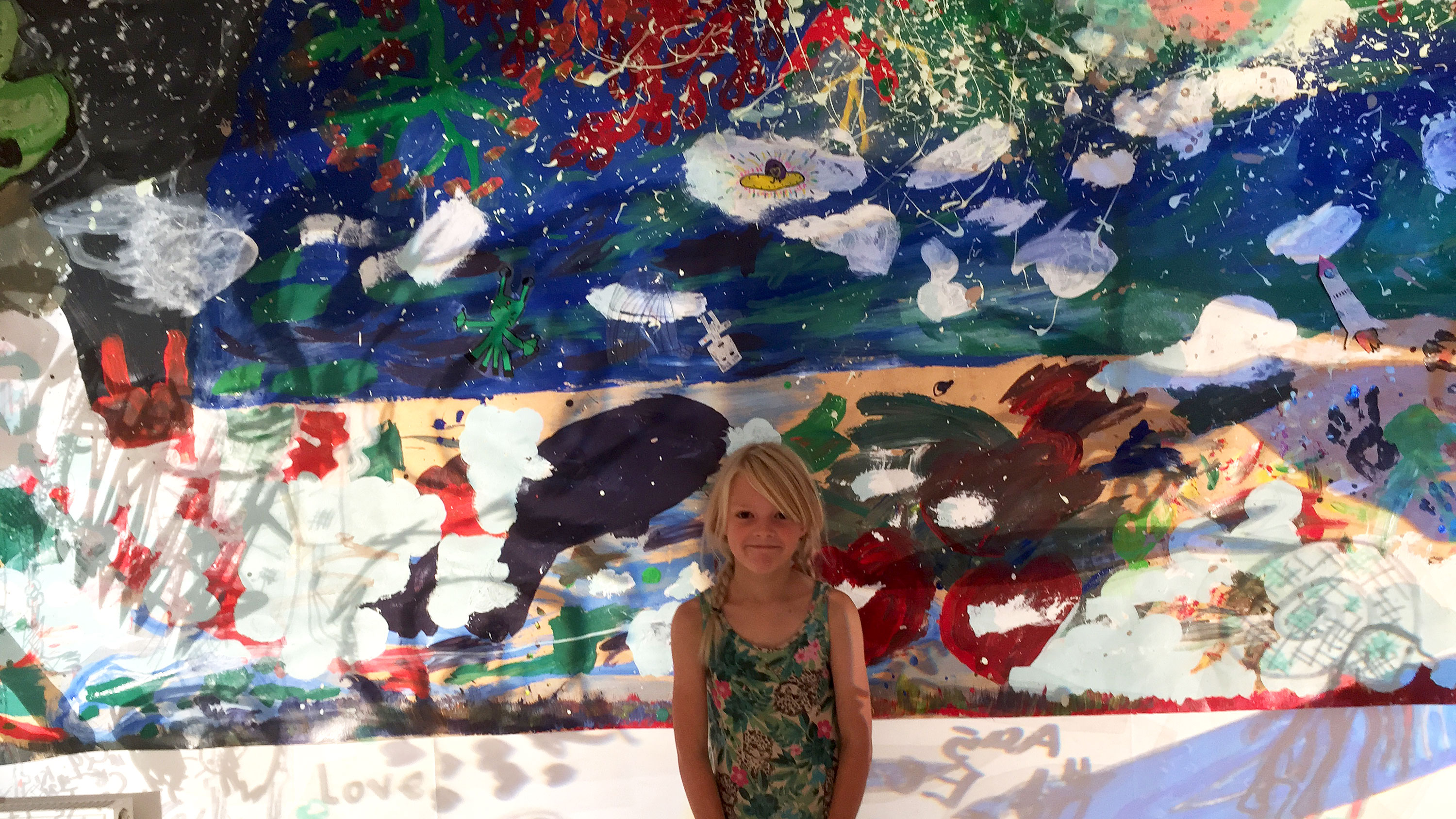 foto av barn med blont hår som står framför en vägg med en stor färgglad målning