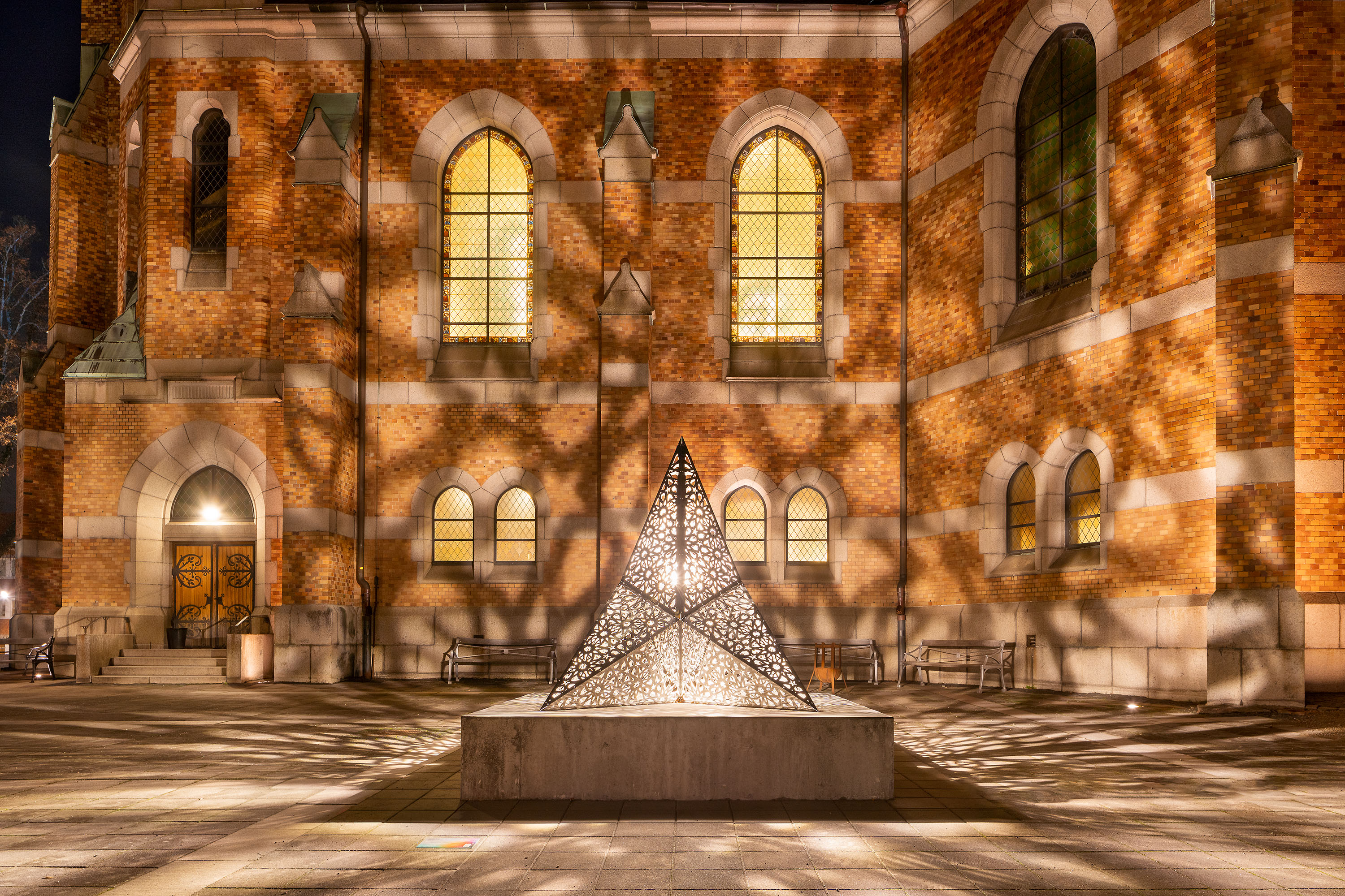 Ljusskulptur i metall med utstansade mönster som kastar mönster i form av skuggor mot en fasad av en kyrka