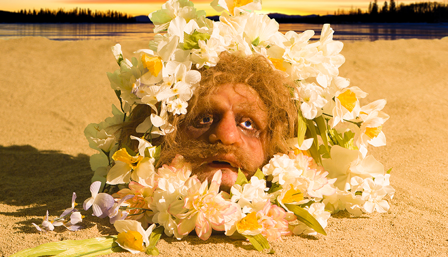 Installation av konstnärerna Nathaniel Mellors och Erkka Nissinen. Mans huvud som sticker ut ur sand omgiven av blommor.