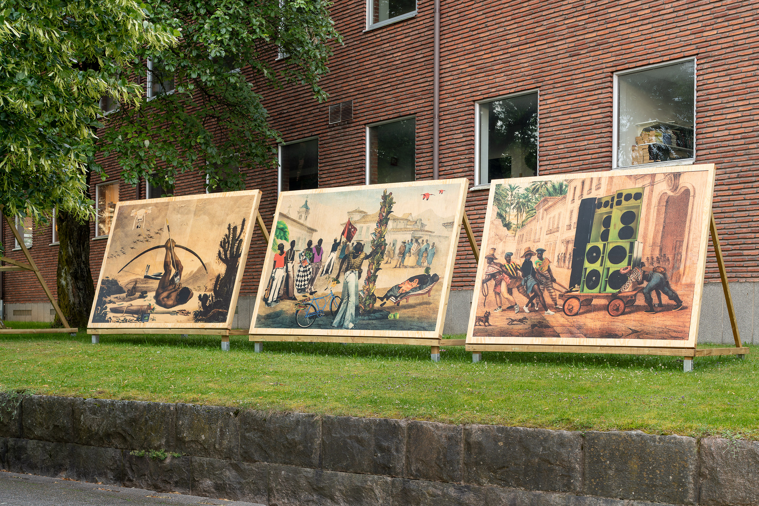 Bilder på plakat är uppställda i gräset framför en byggnad.