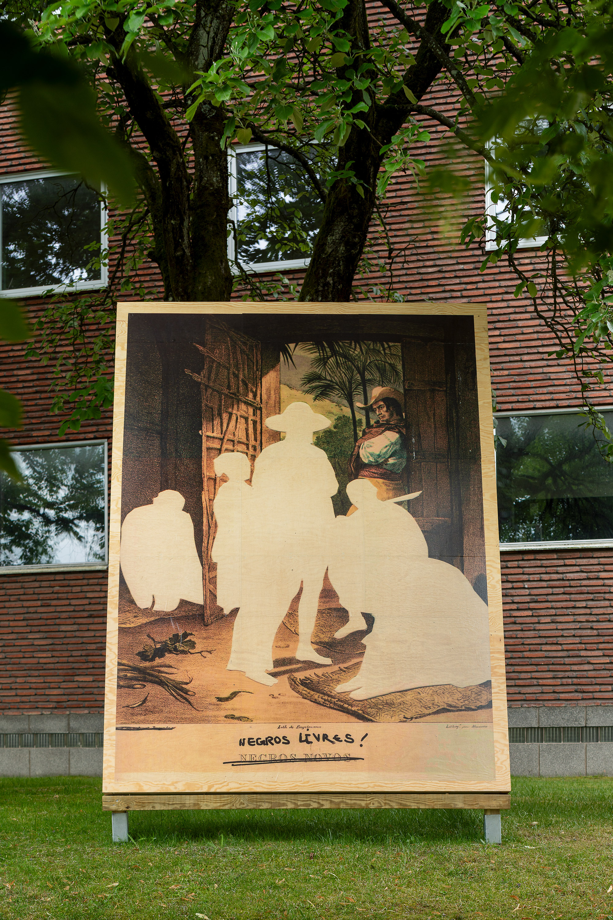 En bild på ett plakat står i gräset framför en byggnad.
