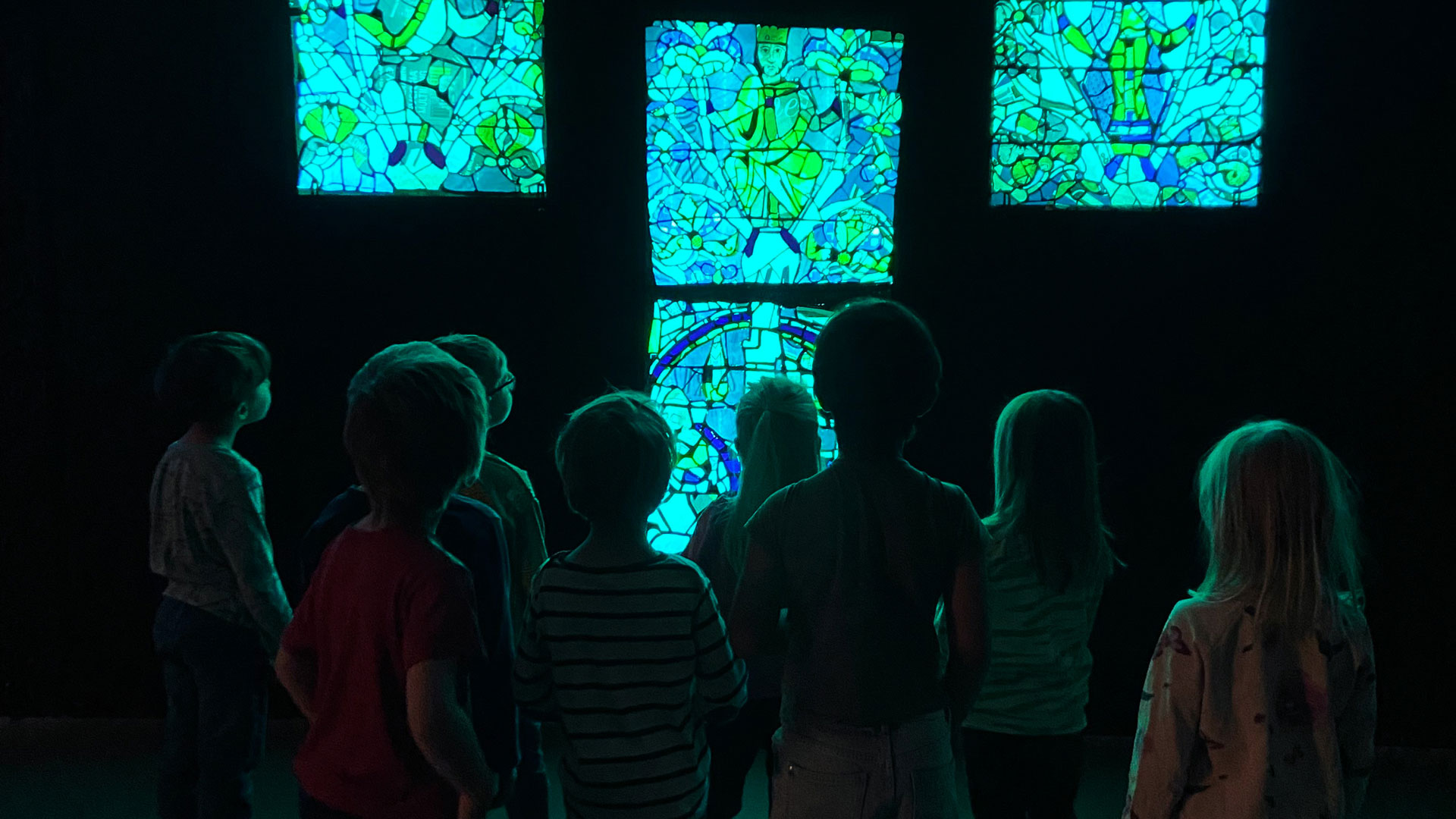 barn i 5-årsåldern står i ett mörkt rum och tittar upp mot ett upplyst konstverk som föreställer ljuslådor med mosaik med olika motiv