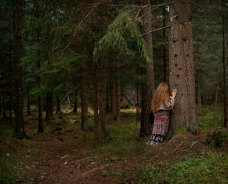 foto föreställande djup skog och en kvinnogestalt i långt hår som lutar pannan mot ett stort träd